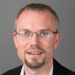 Man with short hair and gotee wearing glasses, black suit, white dress shirt in front of grey background