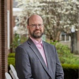 Man with beard wearing grey suit coat and pink button up shirt on porch of Old Centre